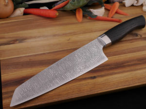Hand Forged Damascus Kitchen knife in the style of a Santoku. It has a Moor Oak handle. Lays of a wooden cutting board. With vegetables in the background