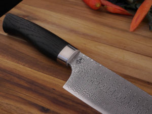 Hand Forged Damascus Kitchen knife in the style of a Santoku. It has a Moor Oak handle. Lays of a wooden cutting board. With vegetables in the background