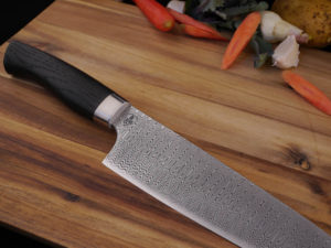 Hand Forged Damascus Kitchen knife in the style of a Santoku. It has a Moor Oak handle. Lays of a wooden cutting board. With vegetables in the background