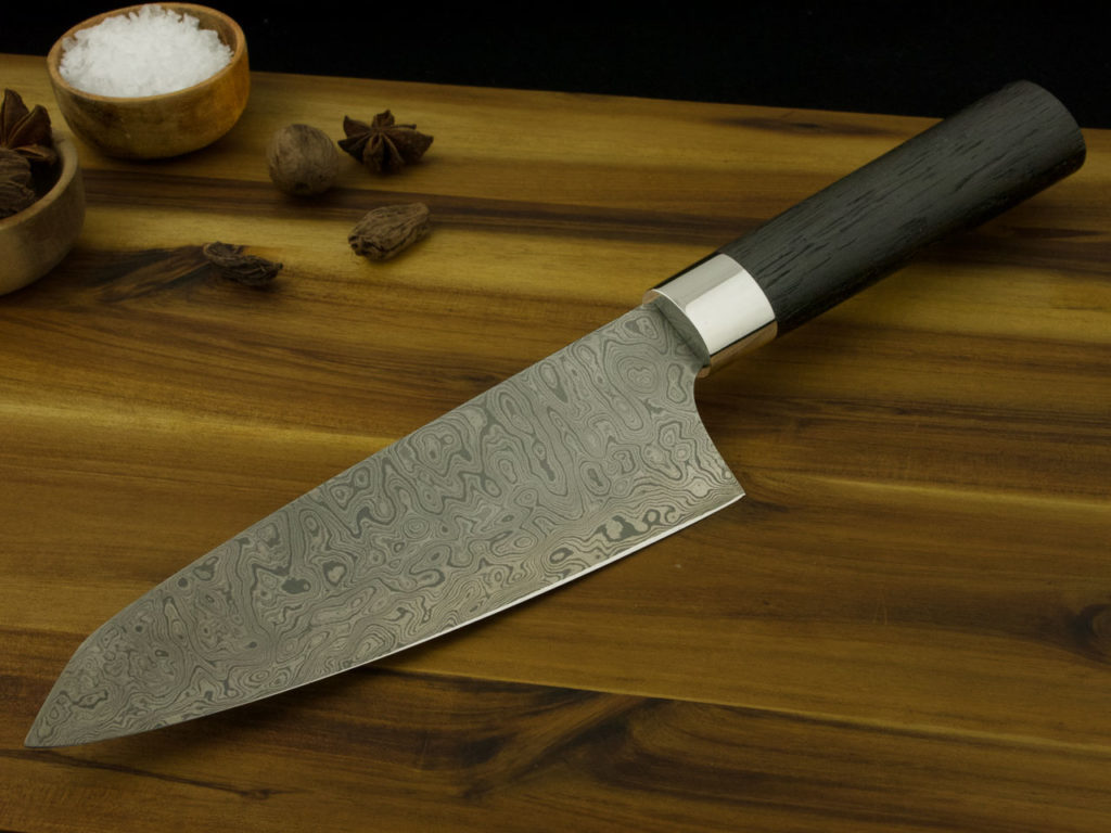 K021-5 Ein maßgefertigtes Santoku-Küchenmesser mit einer Klinge aus Damaszenerstahl und einem Griff aus Mooreiche liegt auf einem Holzschneidebret.