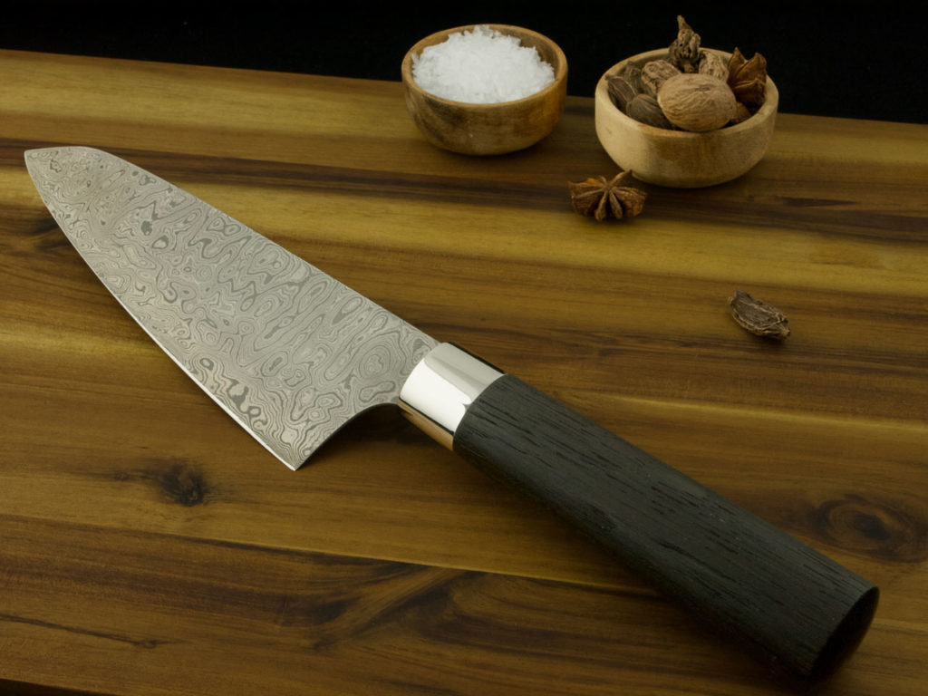 K021-6 Custom Santoku kitchen knife with a Damascus steal blade and handle from Bog Oak lays on a wooden cutting board.