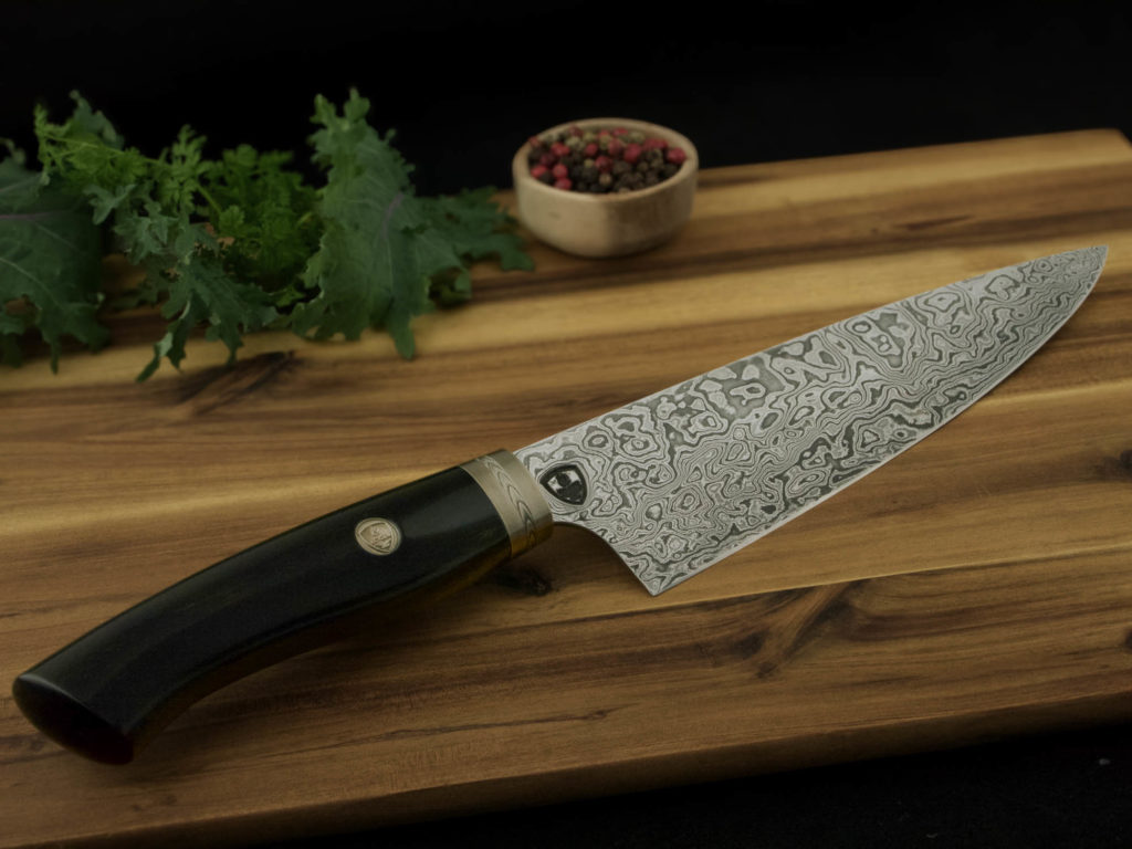 K022-1 professionales Kochmesser aus Damszenerstahl. Griff aus Ebenholz mit handgraviertem Backe aus Neusilber. Das Messer liegt auf einem Holzschniedbrett