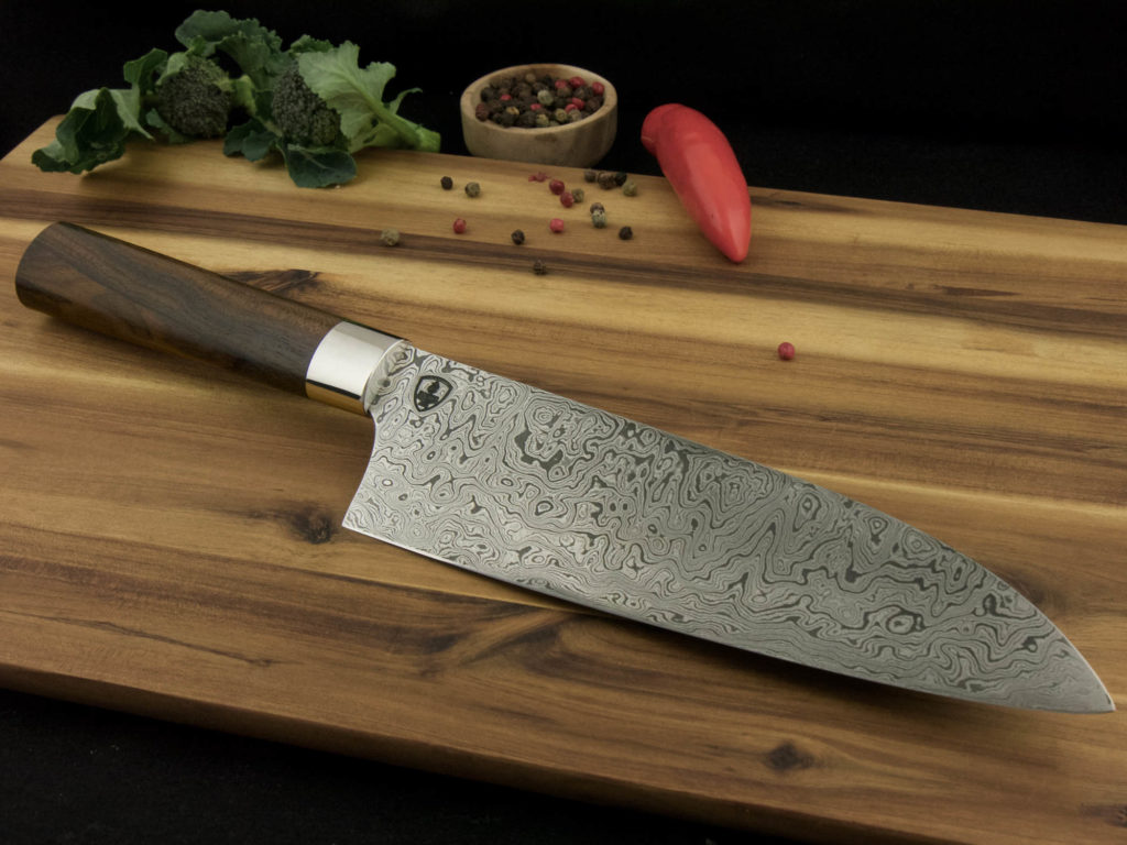 K023-1 Custom Santoku kitchen knife with a Damascus steal blade and handle from Ziricote lays on a wooden cutting board.