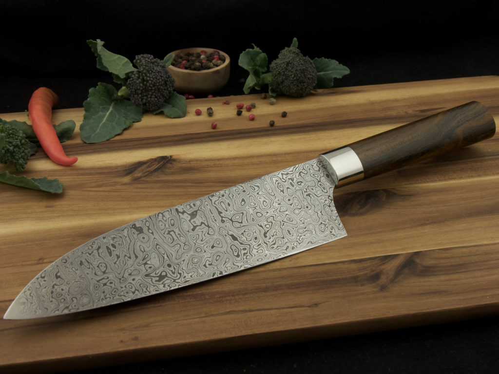 K023-3 Custom Santoku kitchen knife with a Damascus steal blade and handle from Ziricote lays on a wooden cutting board.