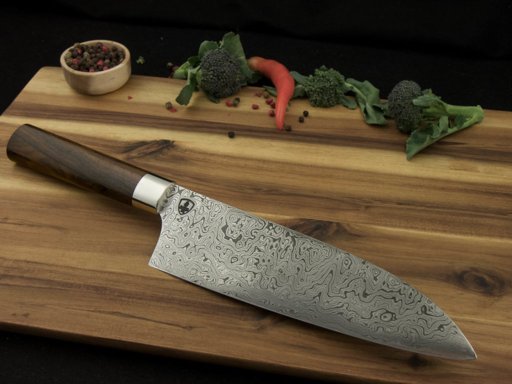 K023-6 Ein maßgefertigtes Santoku-Küchenmesser mit einer Klinge aus Damaszenerstahl und einem Griff aus Ziricote liegt auf einem Holzschneidebret.